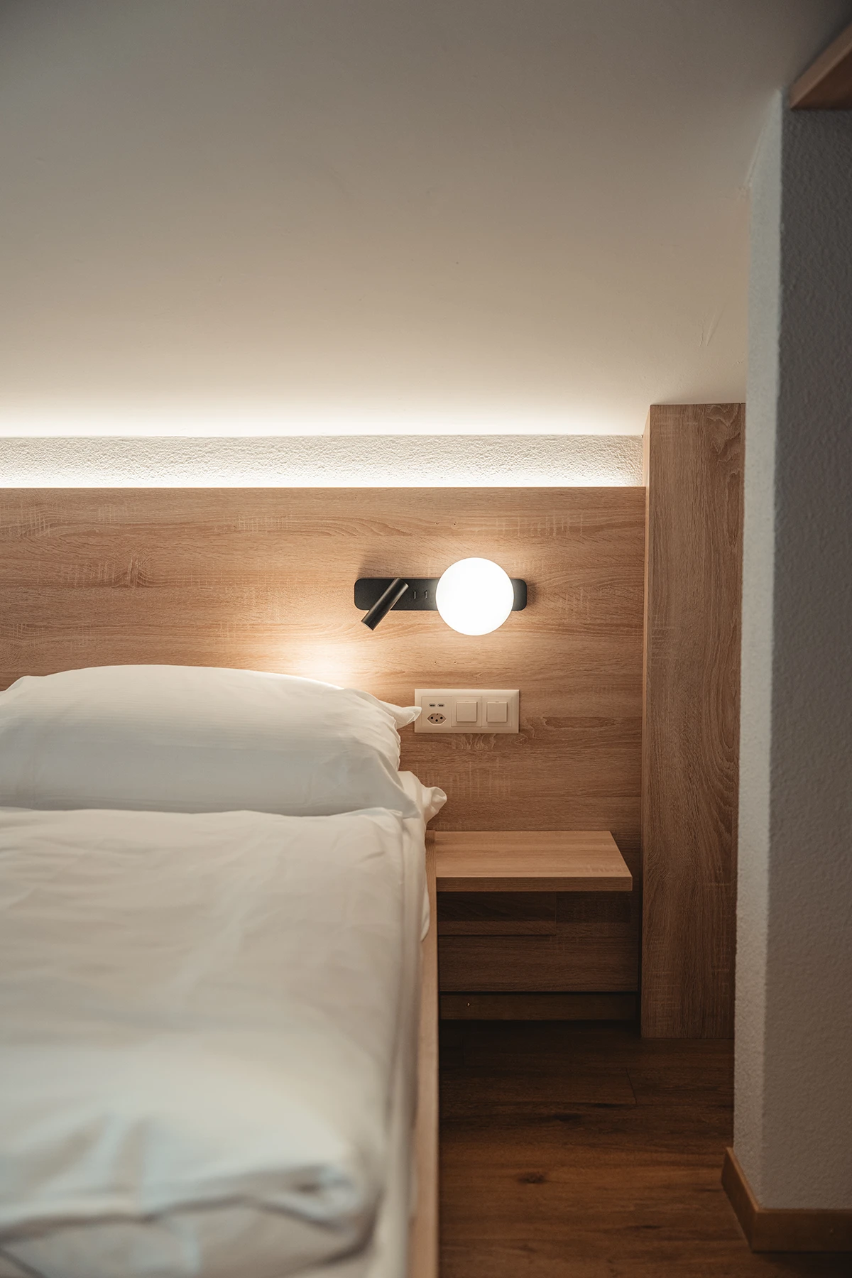 Bett mit weisser Bettwäsche, Holzkopfteil, Leselampe und Nachttisch in einem Zimmer im Matterhorn Inn in Täsch