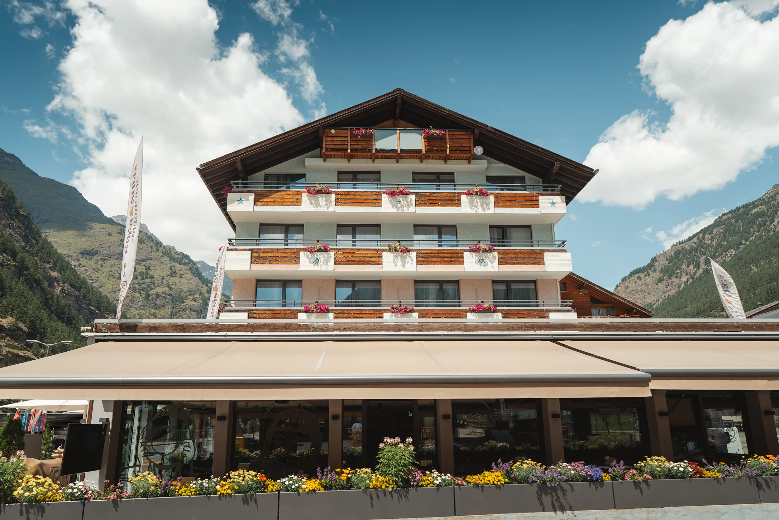 Aussenansicht des Matterhorn Inn in Täsch mit Holzbalkonen, Blumenkästen und Restaurantterrasse vor Bergkulisse
