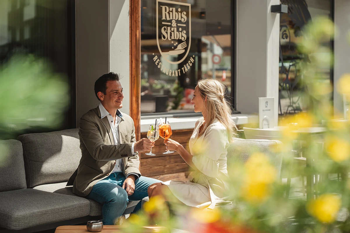Sonnige Terrasse des Restaurants Ribis & Stibis in Täsch: Ein Paar geniesst Aperitifs im Außenbereich des Matterhorn Inn