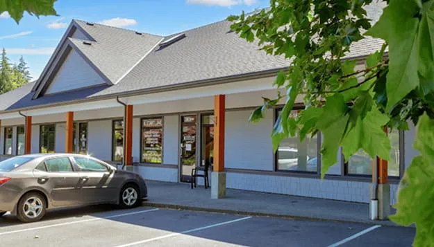 Single-story commercial building with gray siding, large windows, and orange pillars, with a parked dark sedan and green leafy tree in the foreground.