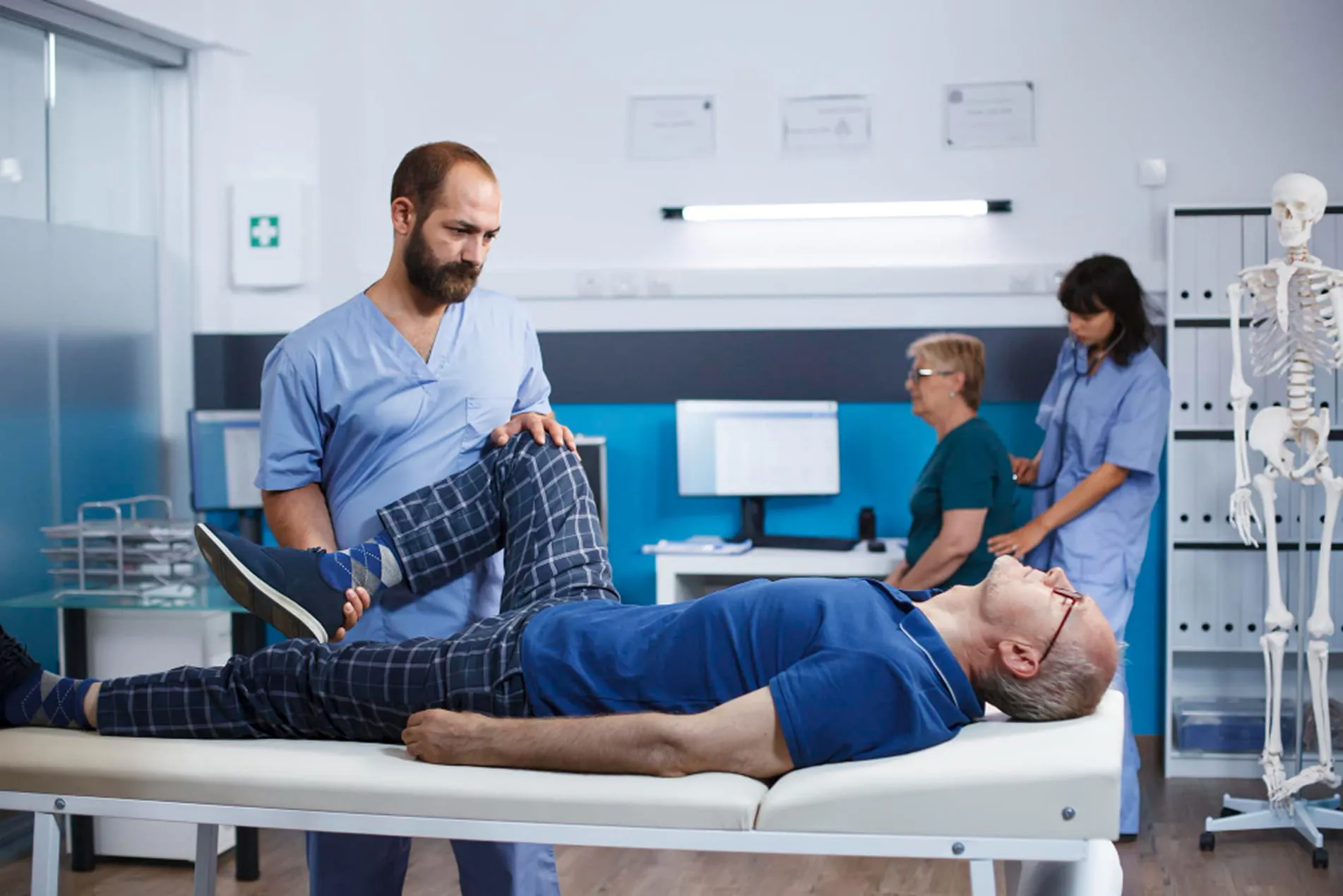 Physical therapist in blue scrubs stretching the leg of a man lying on an examination table in a medical office.