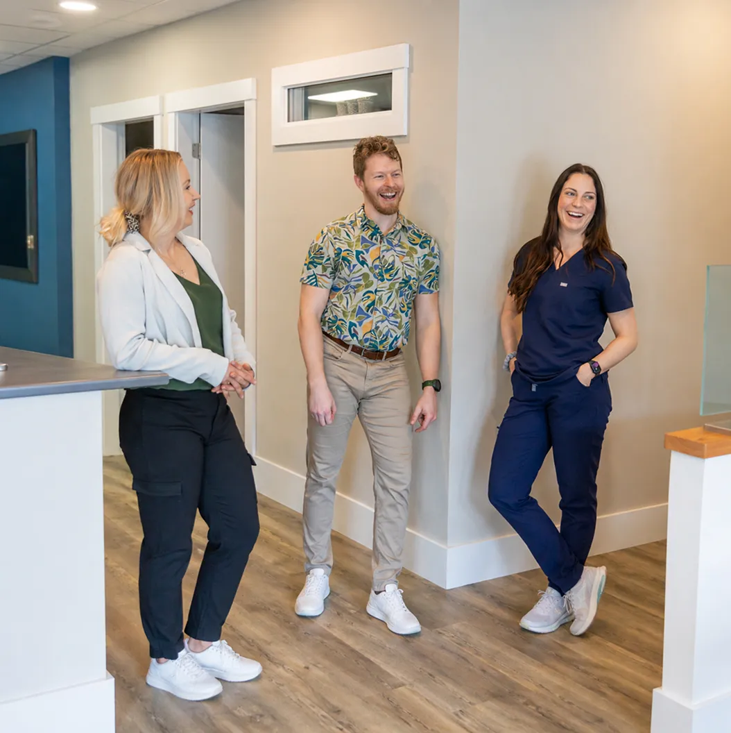 Three smiling professionals standing and chatting in a modern office hallway with wood flooring.