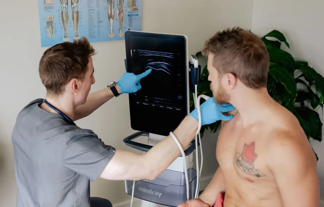 Healthcare professional wearing gloves conducting an ultrasound scan on a shirtless man's neck and pointing at the ultrasound monitor.