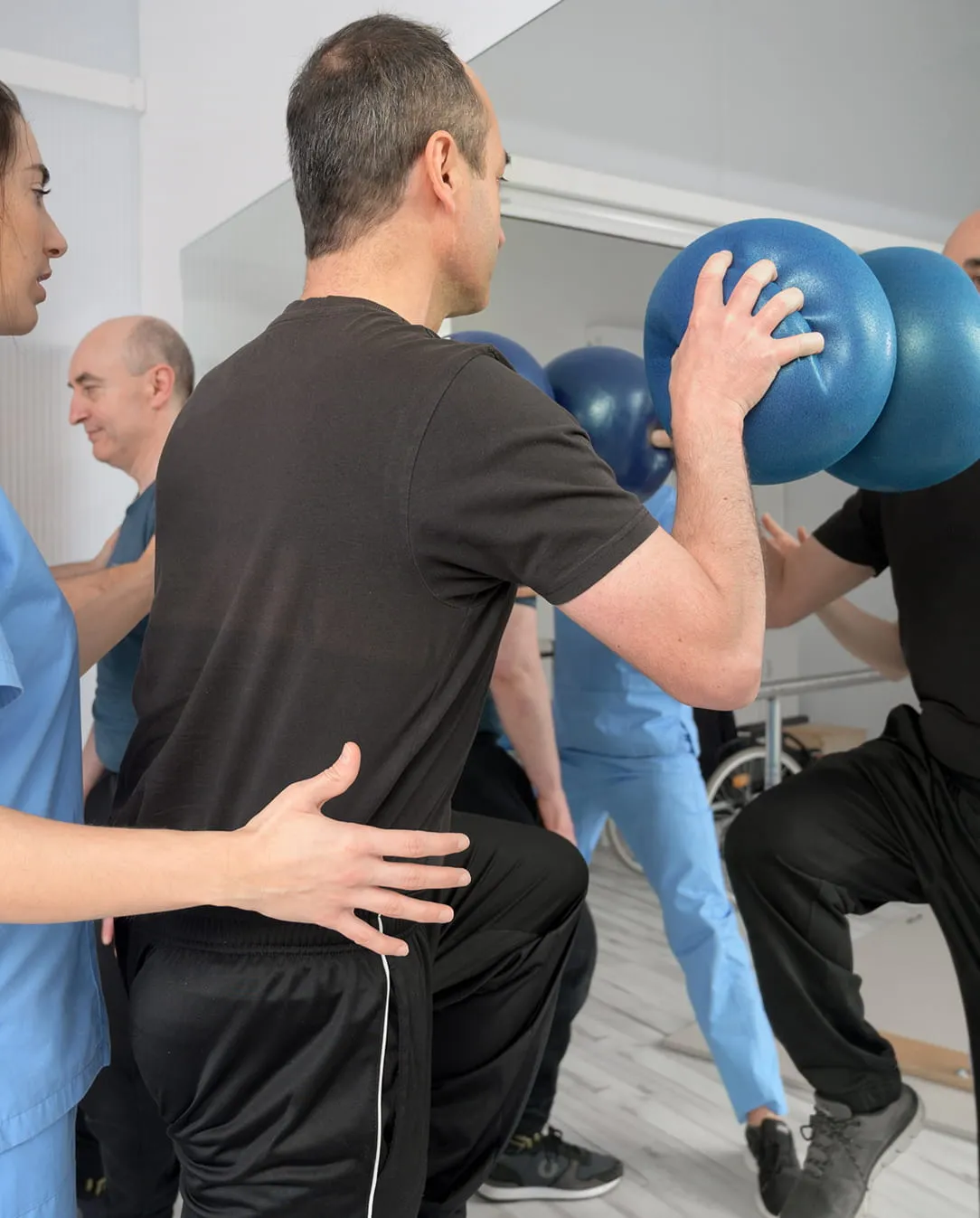 Physical therapist assisting a man holding a blue exercise ball while performing a squat in a gym with mirrors.