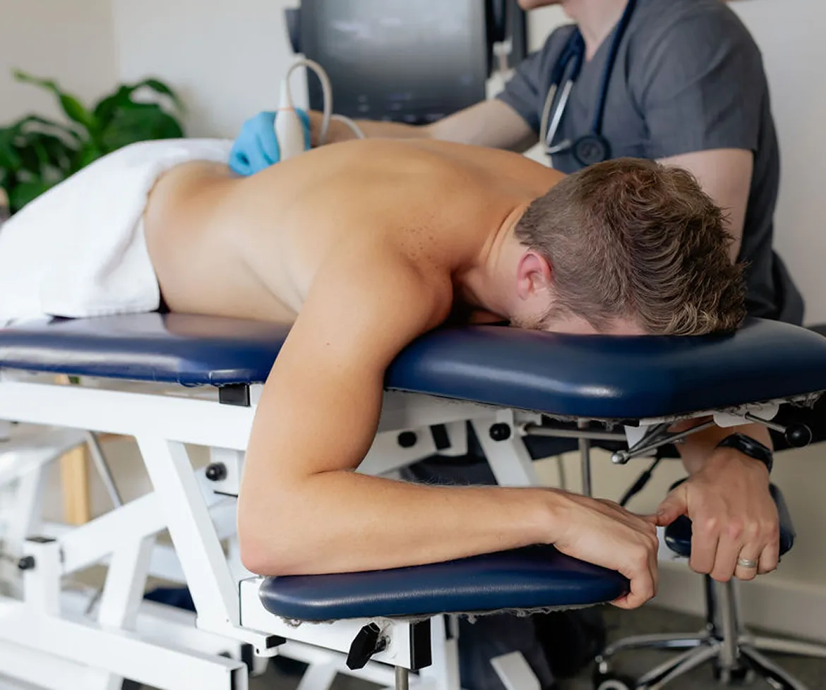 Man lying face down on a medical exam table receiving an ultrasound on his back from a healthcare professional.