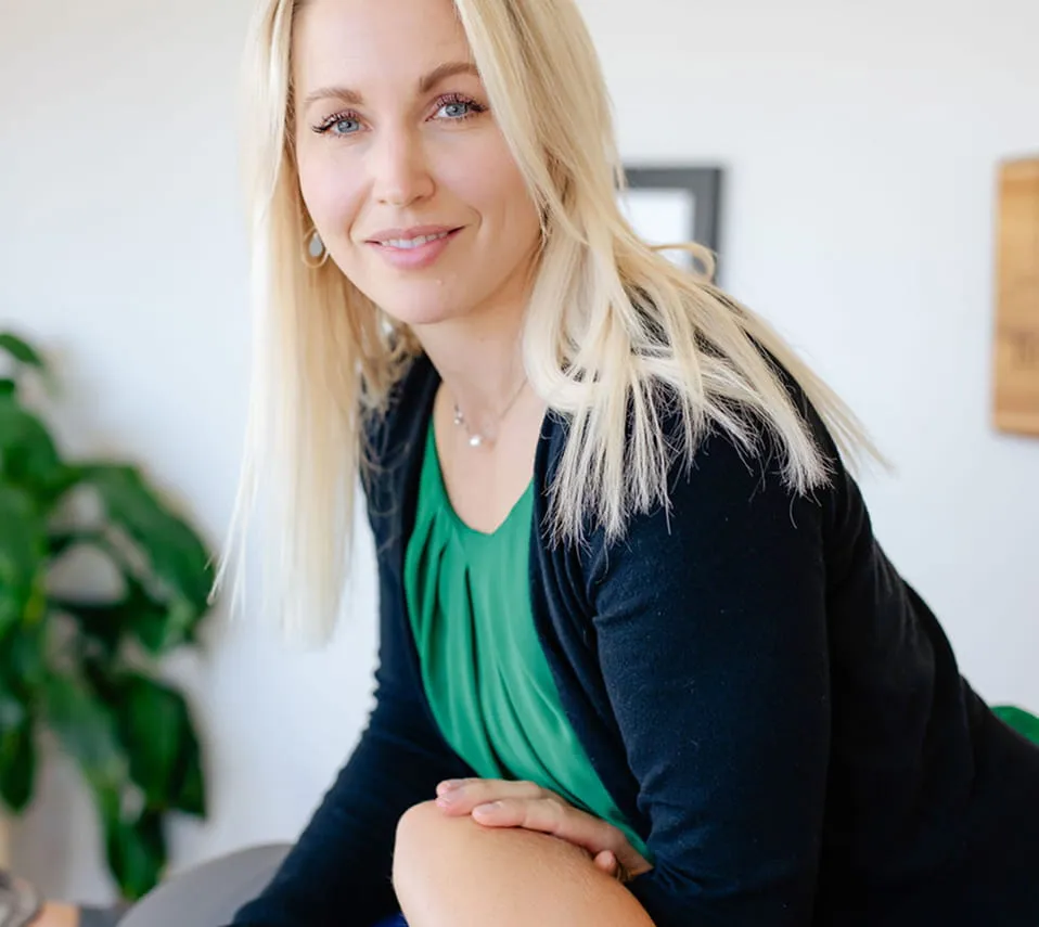 Smiling blonde woman with blue eyes wearing a green top and black cardigan, leaning forward indoors.