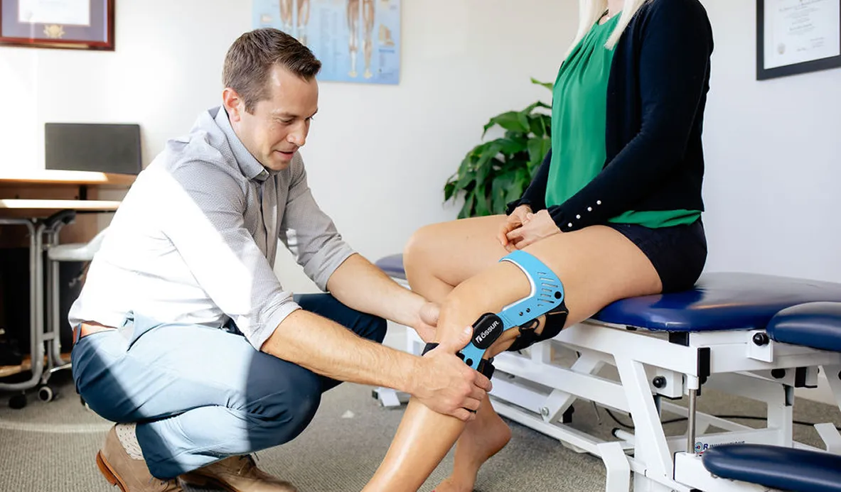 Physical therapist fitting a blue knee brace on a seated woman's leg in a clinical setting.