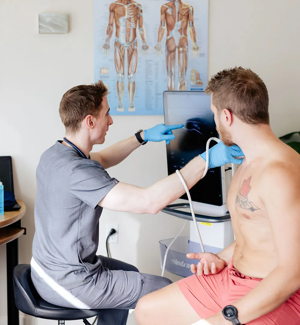 Healthcare professional wearing gloves using an ultrasound device to examine a man's shoulder while showing the scan on a monitor.