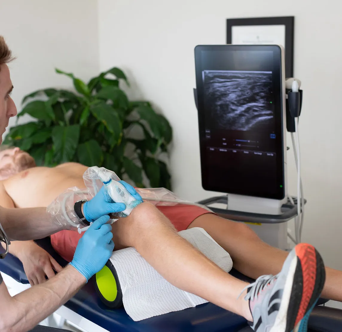 Medical professional performing an ultrasound scan on a patient's knee using a handheld probe connected to a monitor displaying the ultrasound image.