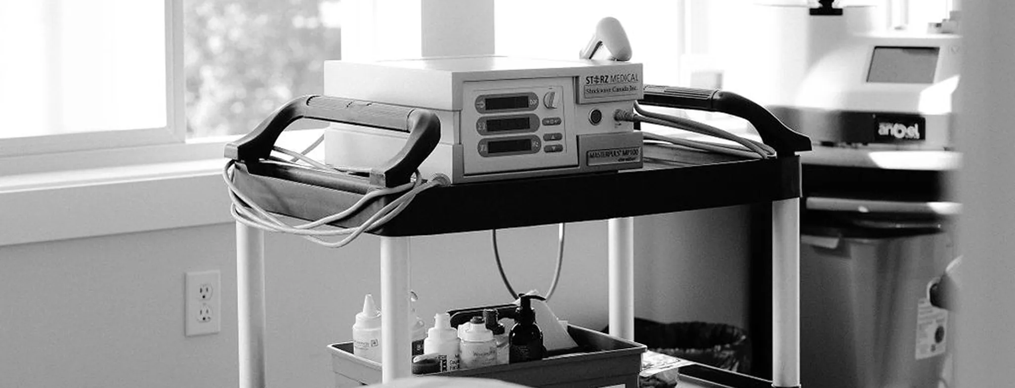 Medical cart with electronic monitoring equipment and various bottles in a clinical room near a window.