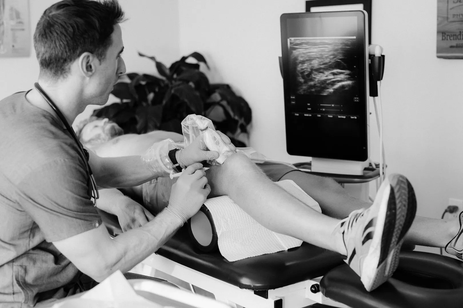 Doctor performs ultrasound-guided injection on patient's knee in medical exam room.
