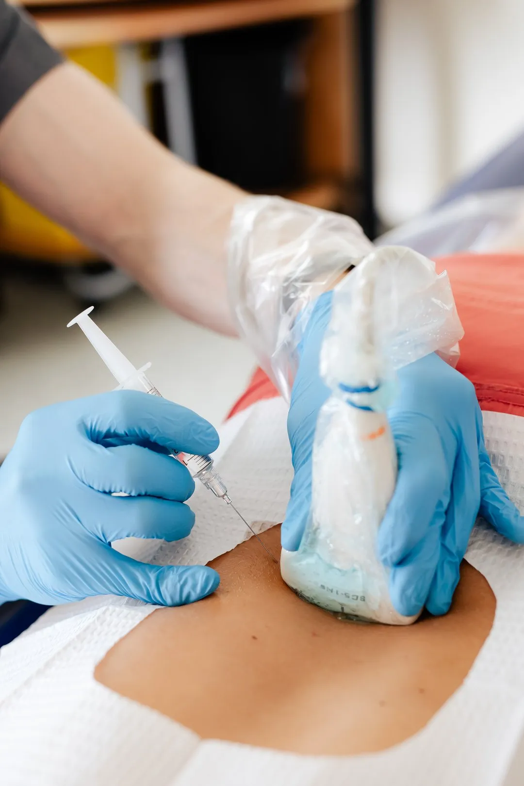 Healthcare worker in blue gloves using ultrasound and needle on patient’s abdomen for injection or biopsy.