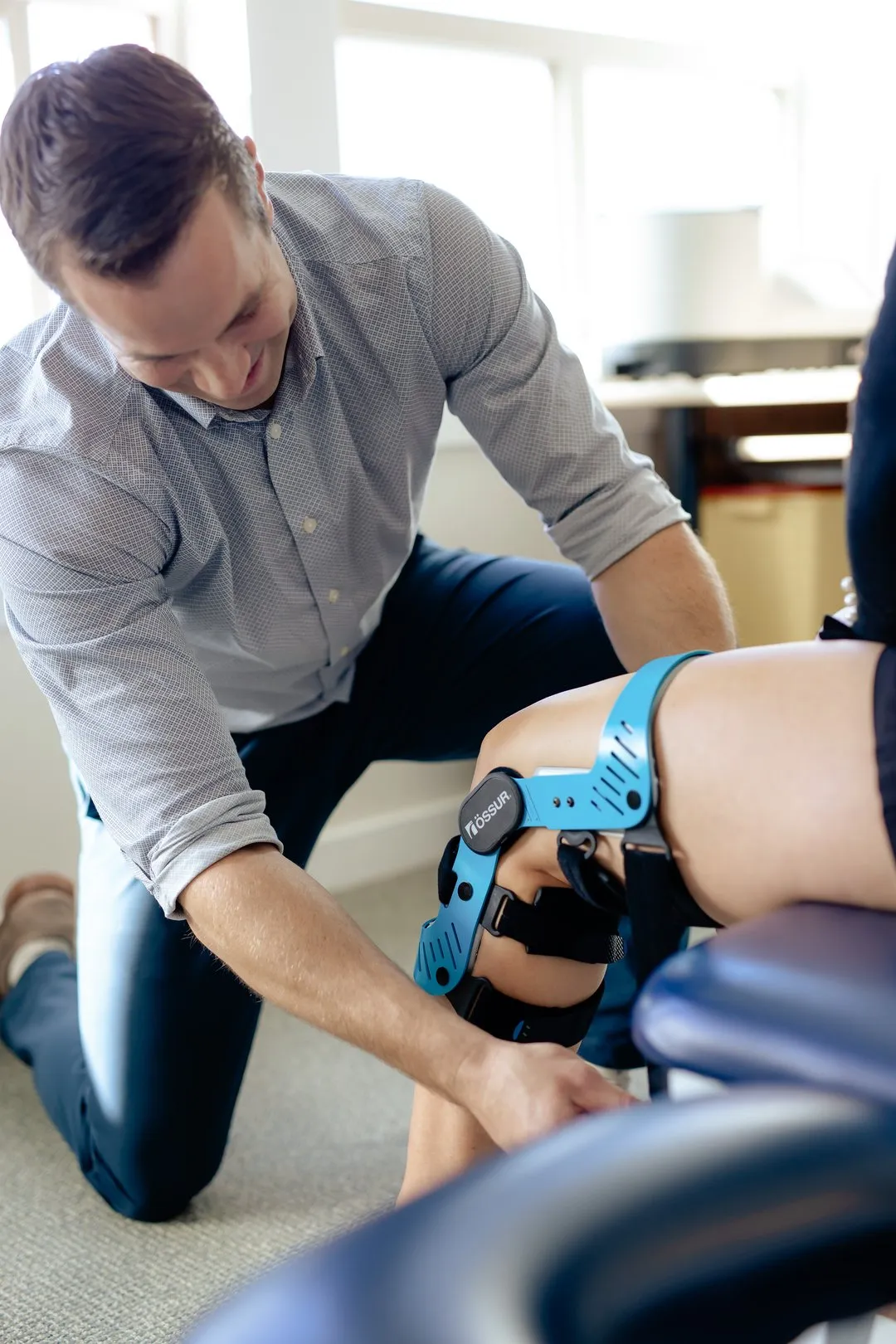 Man fitting a blue knee brace labeled Össur on a seated person's knee in a bright room.