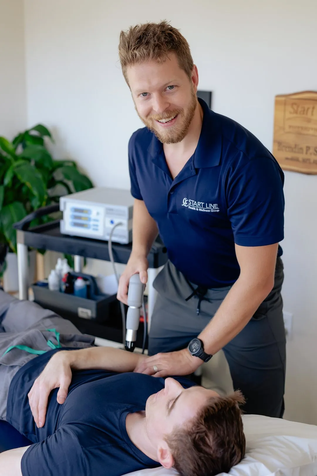 Therapist in navy shirt using a handheld device on a patient's shoulder in a clinic room.