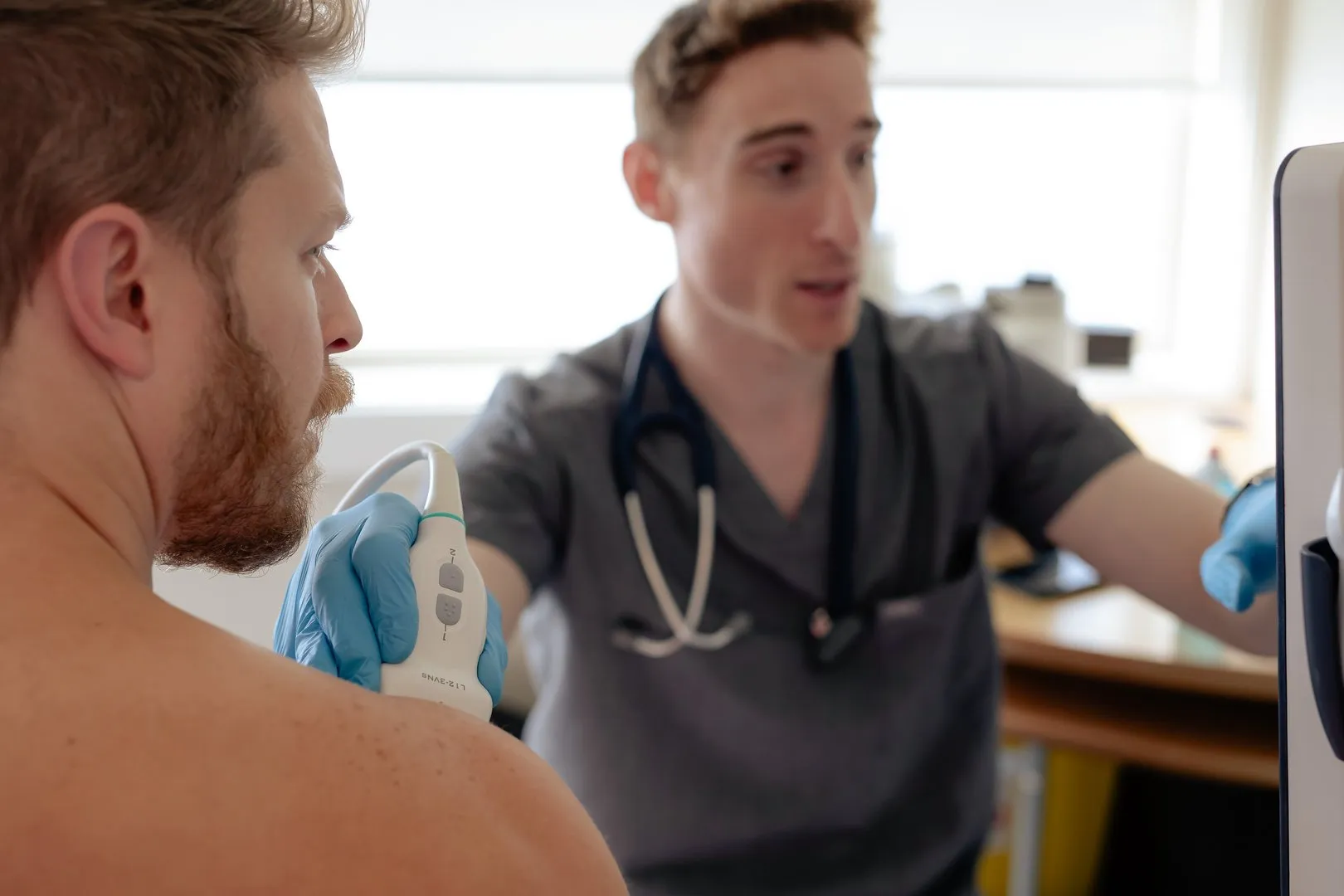Medical professional using ultrasound device on shirtless man's shoulder while viewing monitor.
