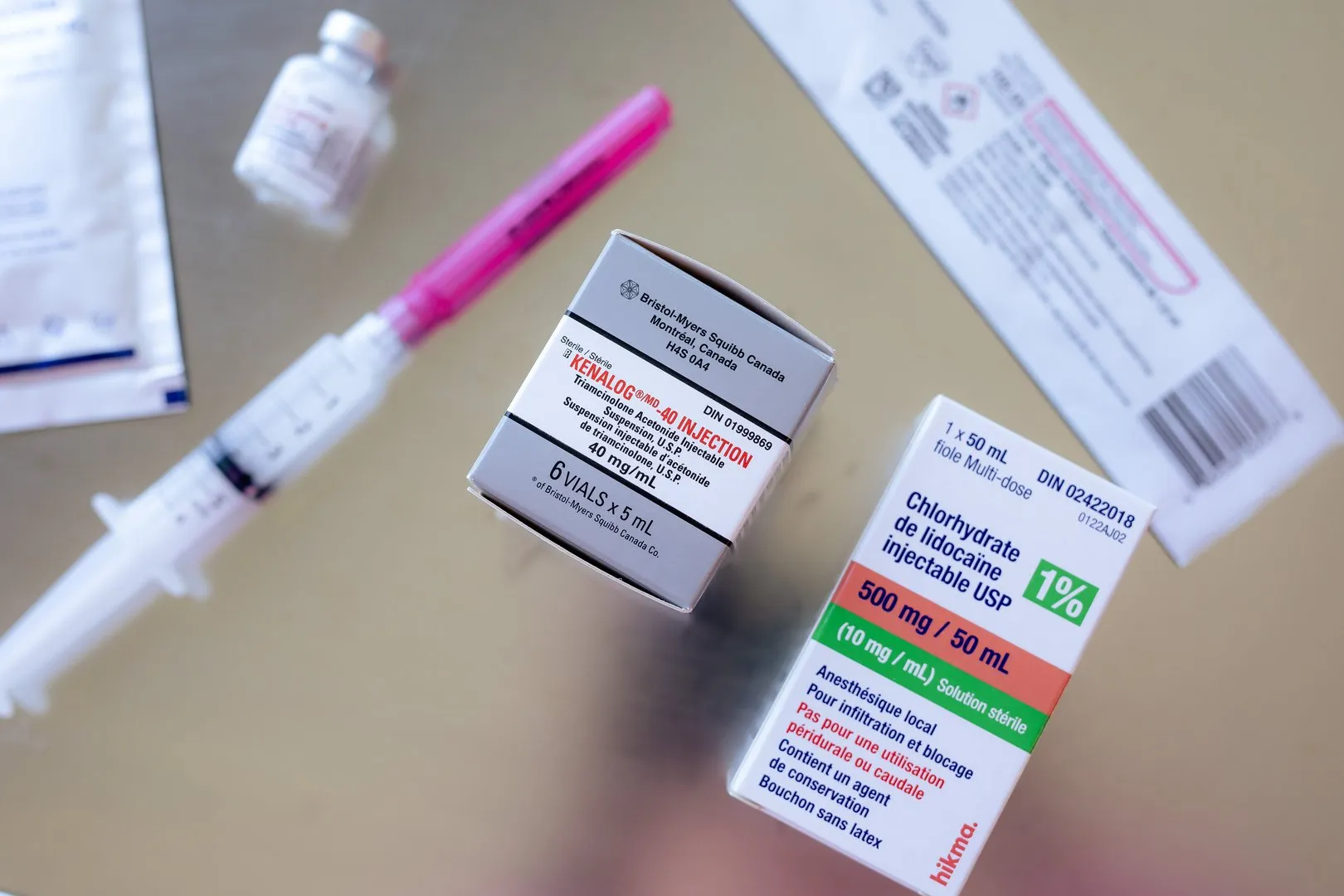 Two medication boxes and a syringe on a reflective surface, including Kenalog and lidocaine injectable.