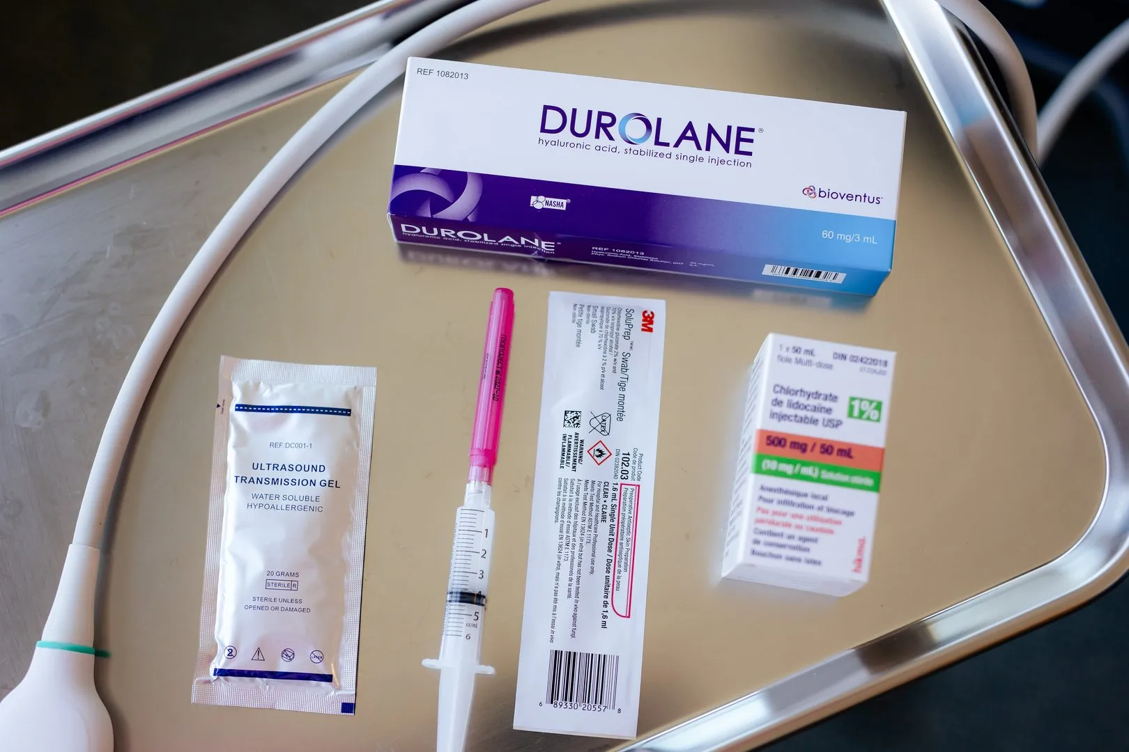 Medical items on metal tray: ultrasound gel, syringe with pink cap, 3M swab, lidocaine vial, and Durolane injection box.