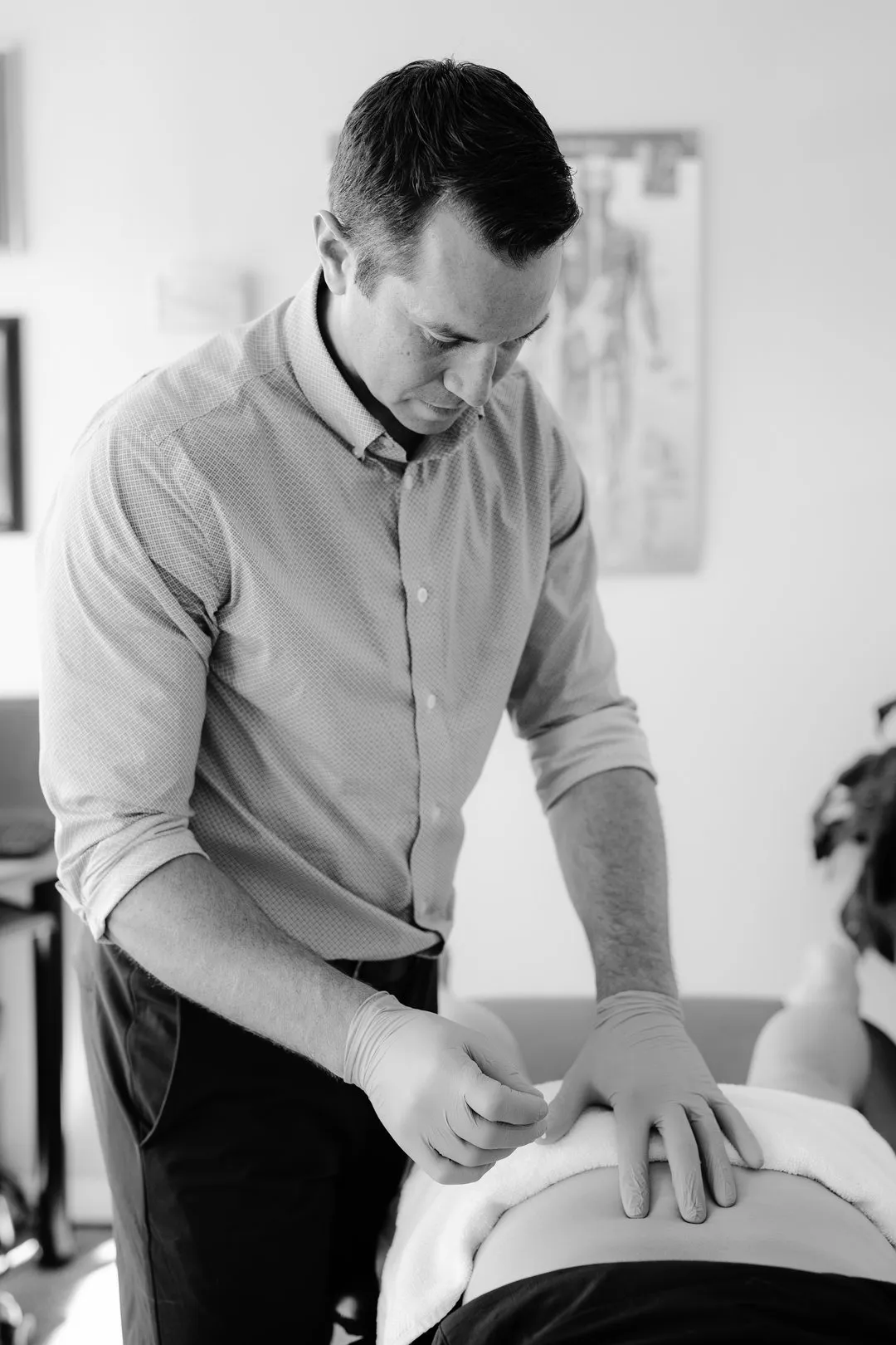 Therapist wearing gloves performs acupuncture on a patient's lower back in a clinical room.