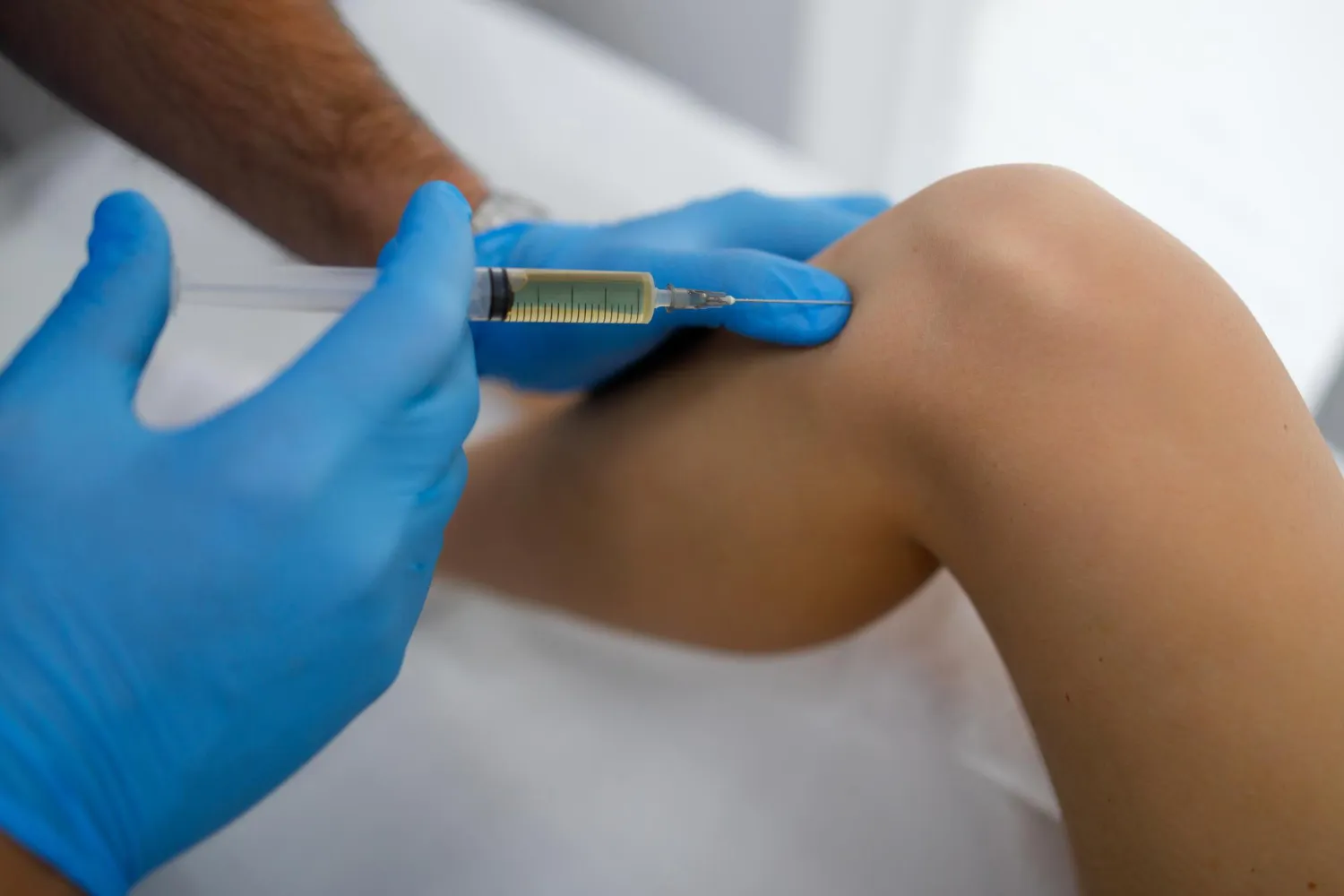 Gloved hands injecting liquid with a syringe into a bent knee joint.