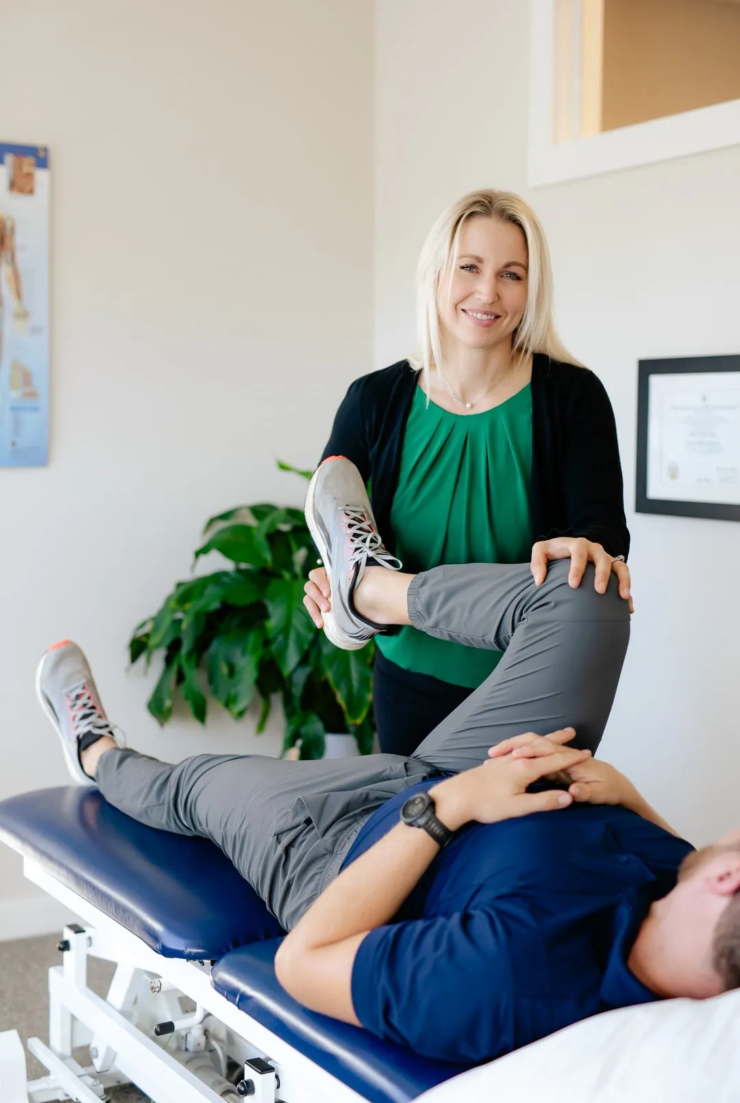 Female physical therapist in green top stretches a man's leg on an exam table in a clinic.