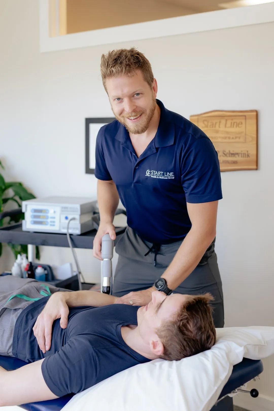 Therapist in navy shirt using a handheld device on a male patient lying on a treatment table.