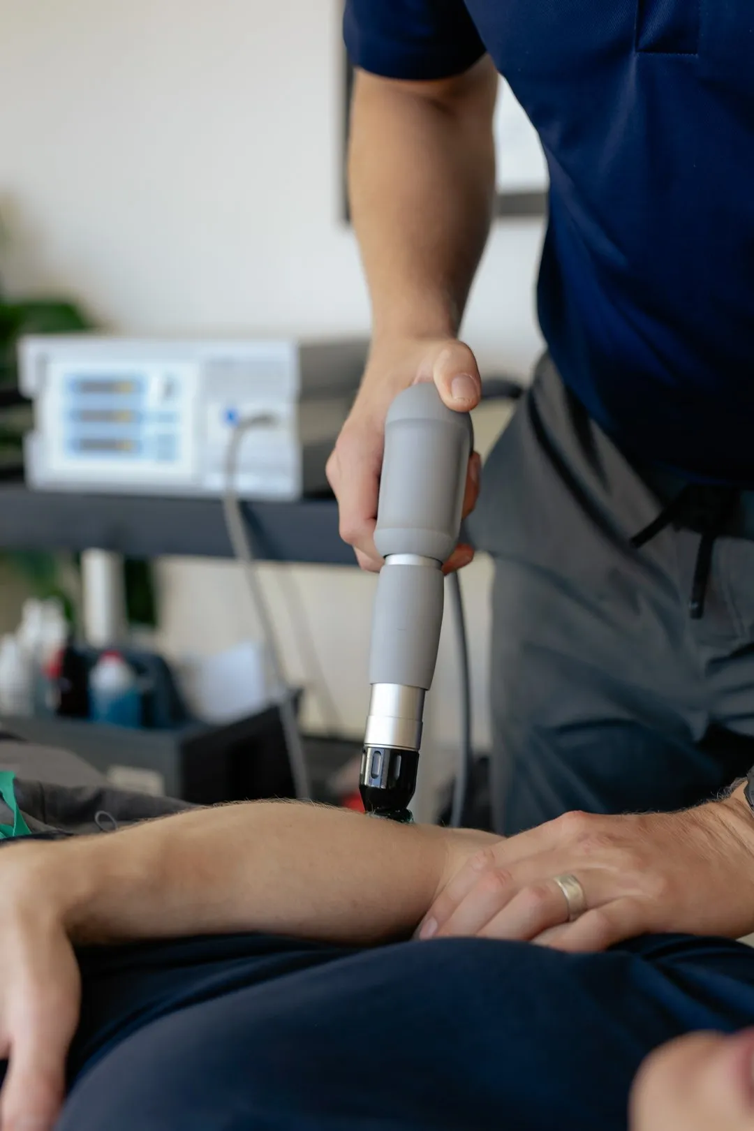 Therapist using a handheld therapy device on a patient's forearm in a clinical setting.