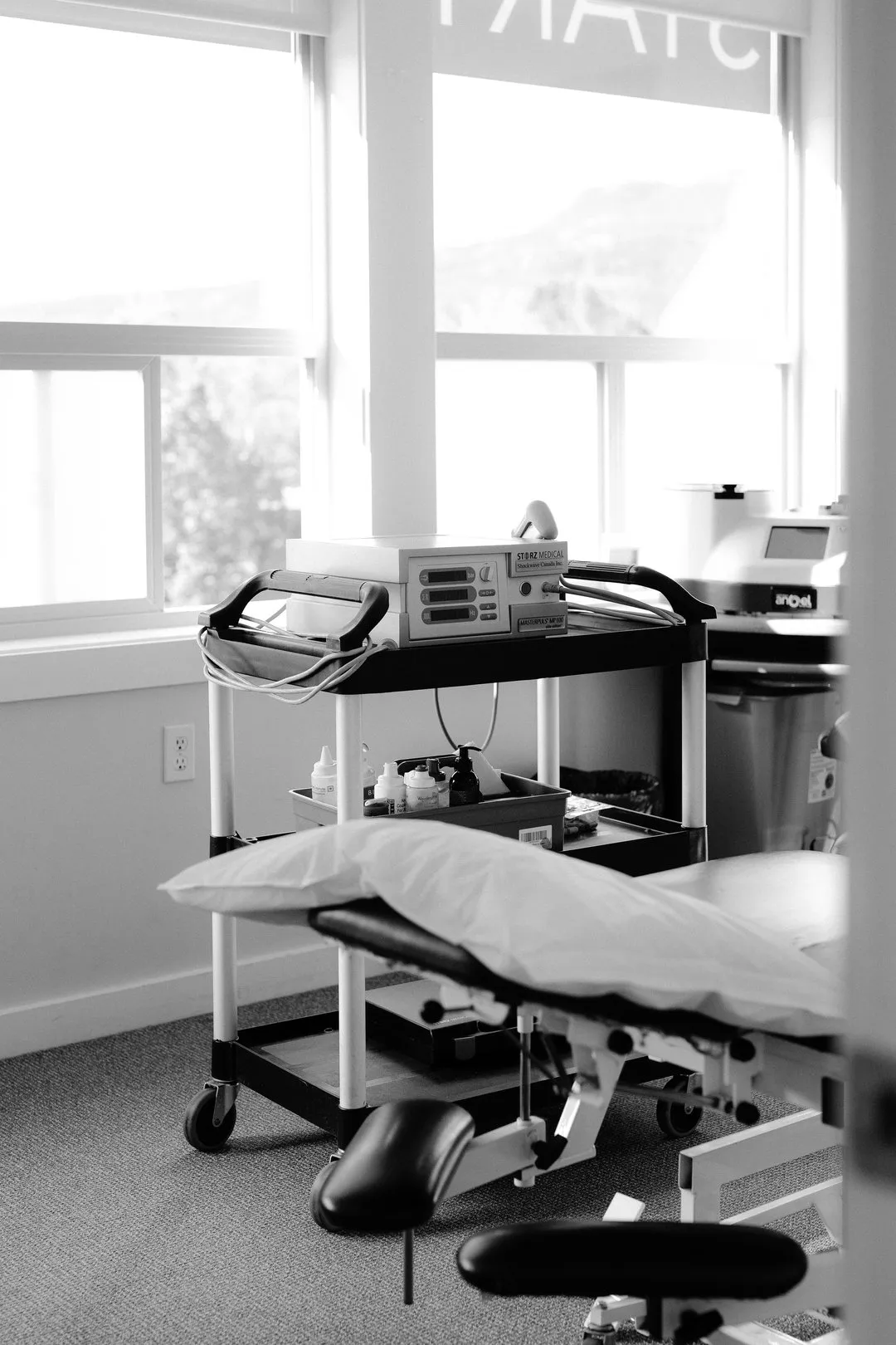 Medical examination room with a padded table, pillow, and equipment cart holding medical devices and bottles.