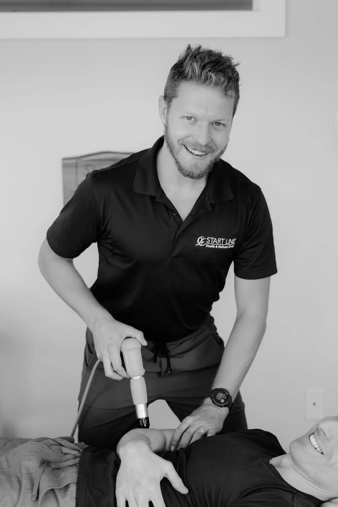 Smiling male therapist in Start Line shirt uses handheld device on a patient's arm while patient lies down.