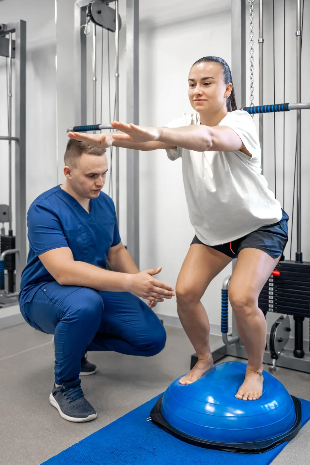 Woman balancing on a blue exercise ball while a trainer in blue scrubs spots her squat form.