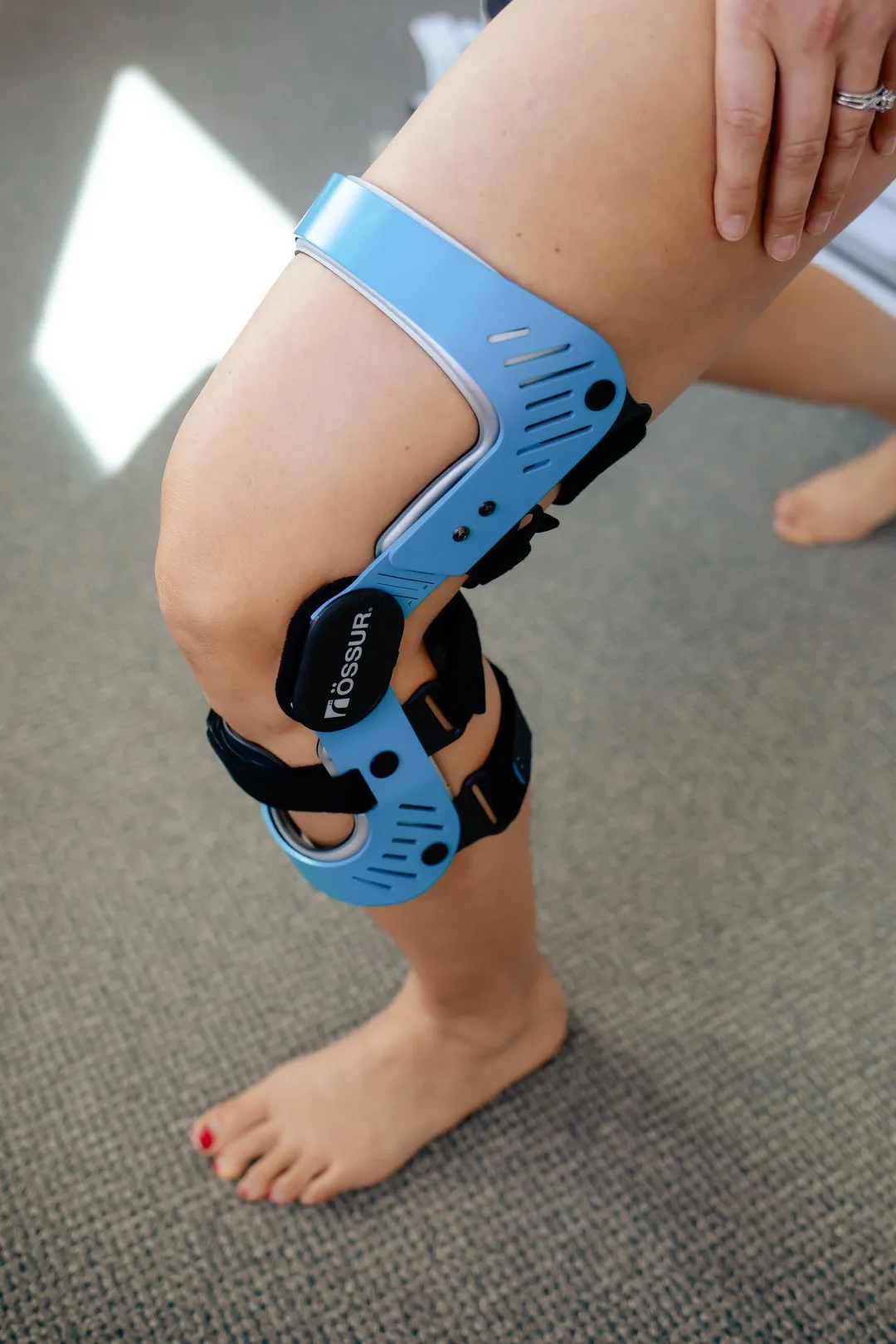 Person wearing a blue Össur knee brace on a bent leg standing on carpeted floor.