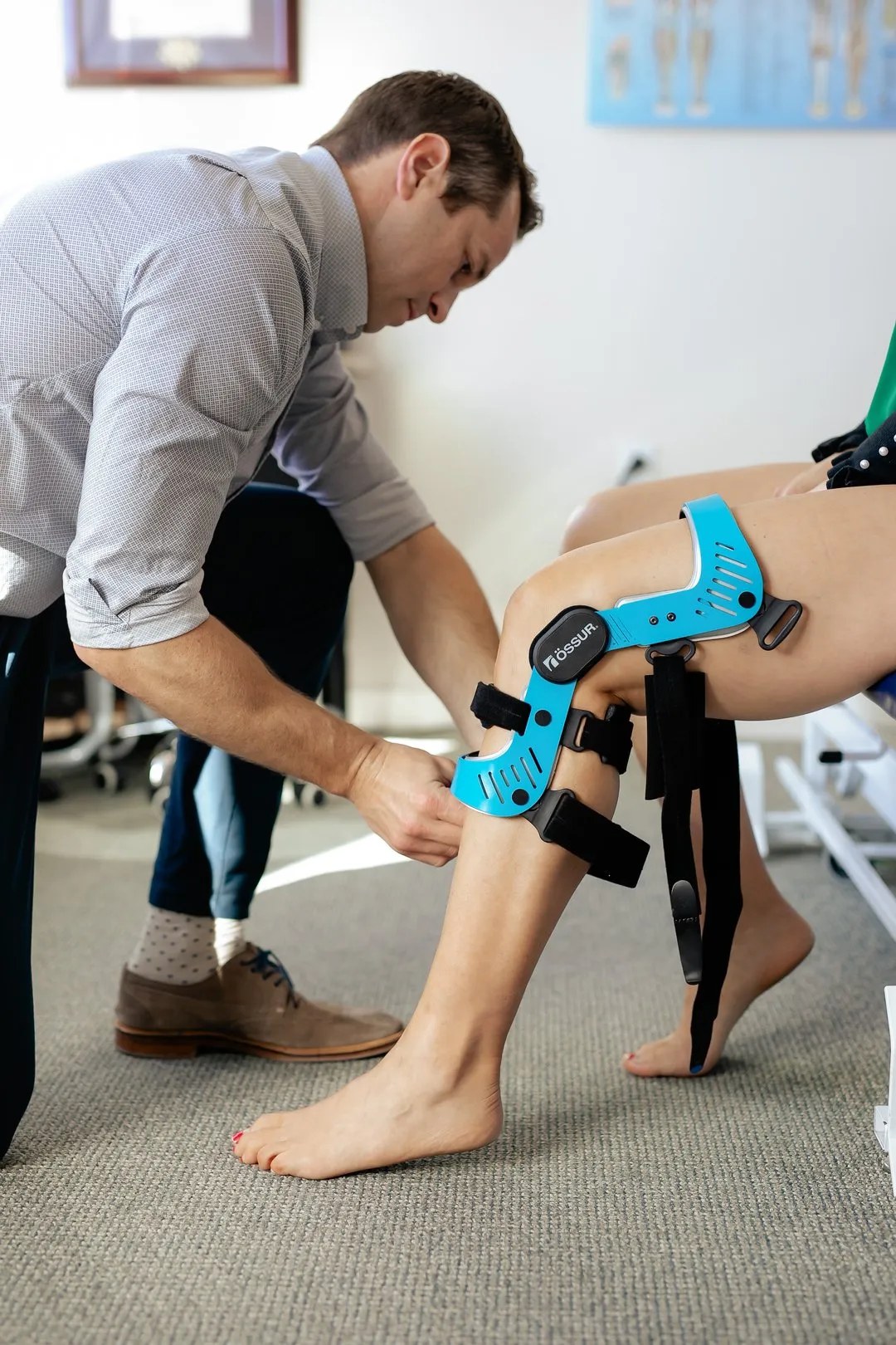 Man fitting blue knee brace with straps on a seated woman's leg in a clinical setting.