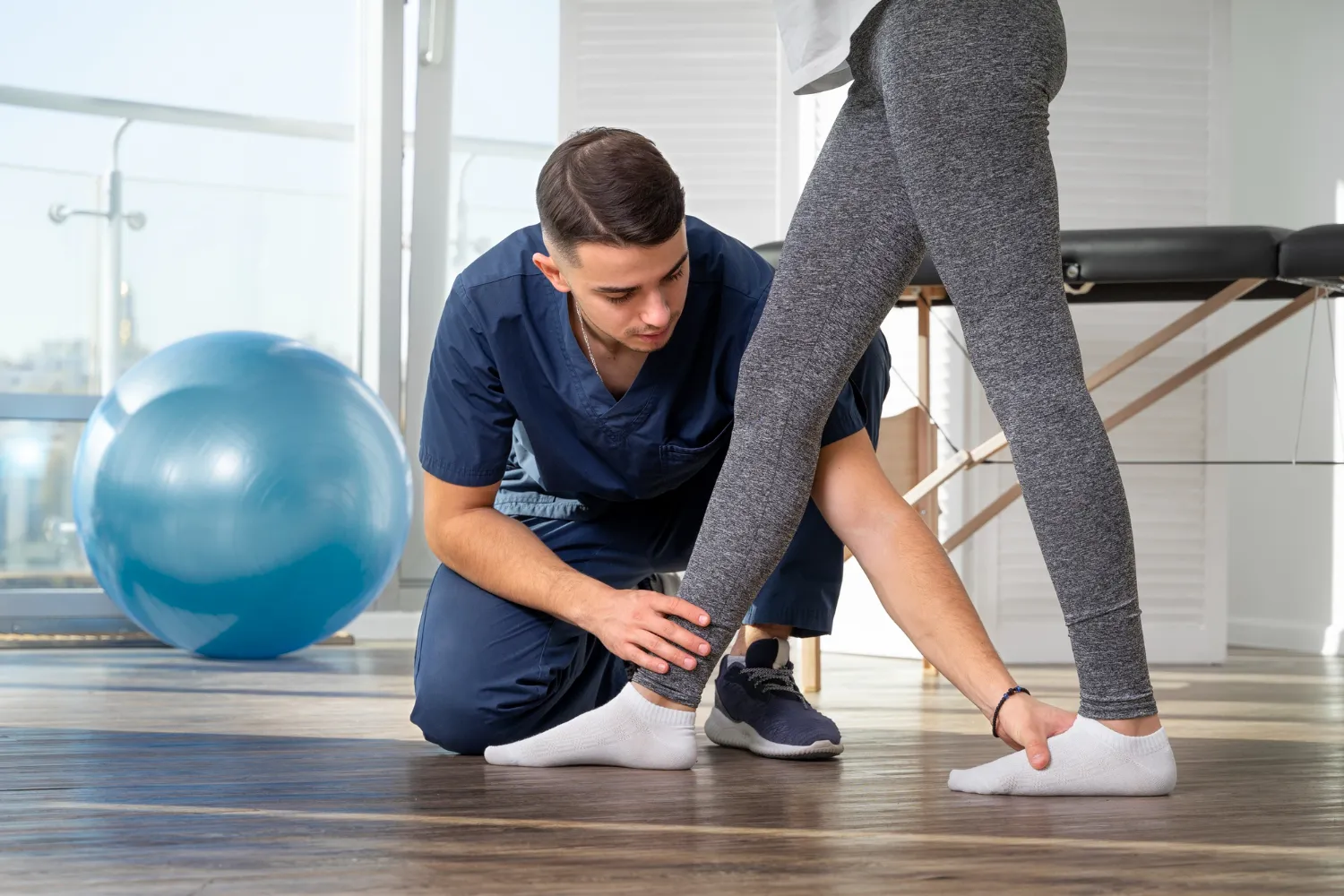 Physiotherapist examining a patient's ankle and calf in a brightly lit therapy room.
