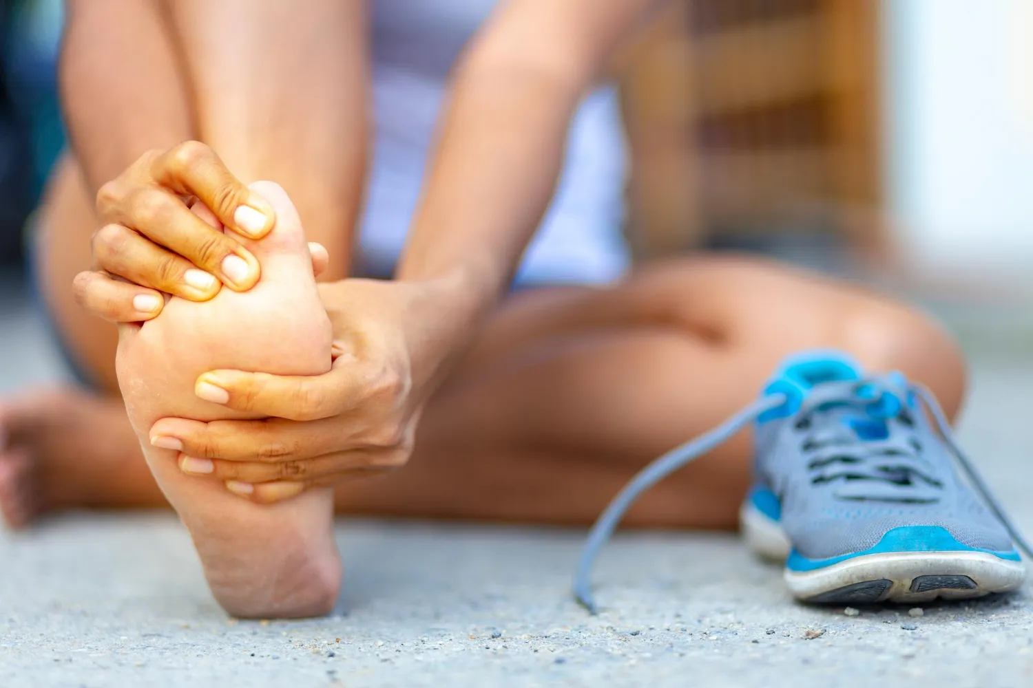 Person sitting on ground holding and rubbing the sole of one foot with a blue sneaker beside them.