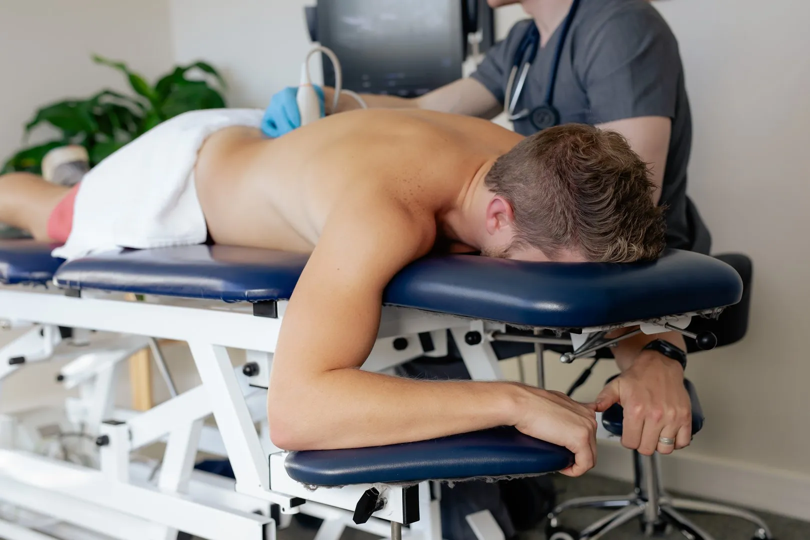Man lying face down on medical table receiving back ultrasound from healthcare professional.