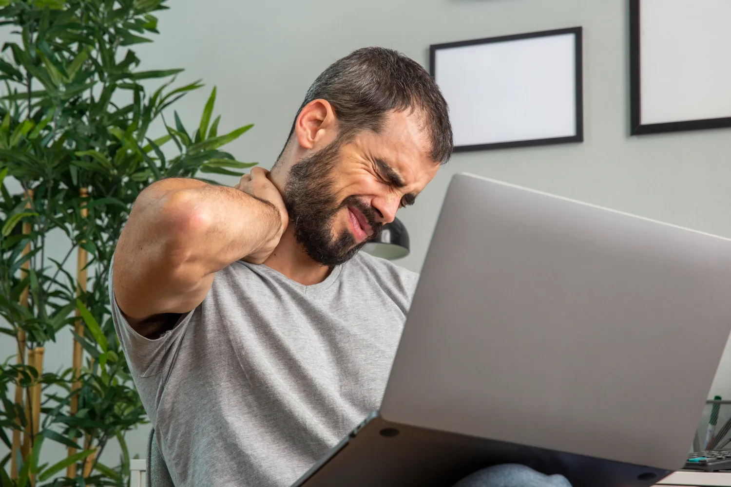 Man in gray shirt holding his neck in pain while using a laptop at home.