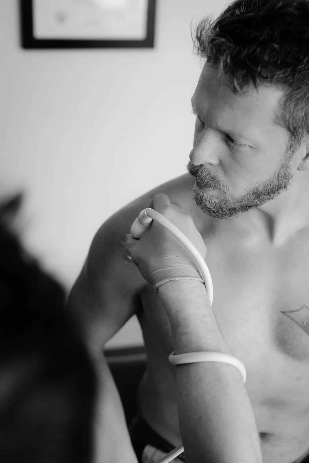 Medical professional using an ultrasound device on a shirtless man's shoulder in a clinical setting.
