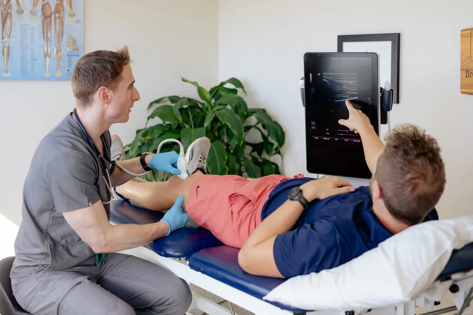 Doctor using ultrasound device on knee of patient lying on exam table pointing at monitor screen.