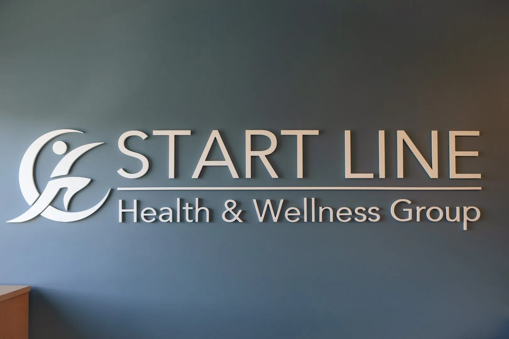 Wall sign of START LINE Health & Wellness Group with a running person logo on a gray wall.