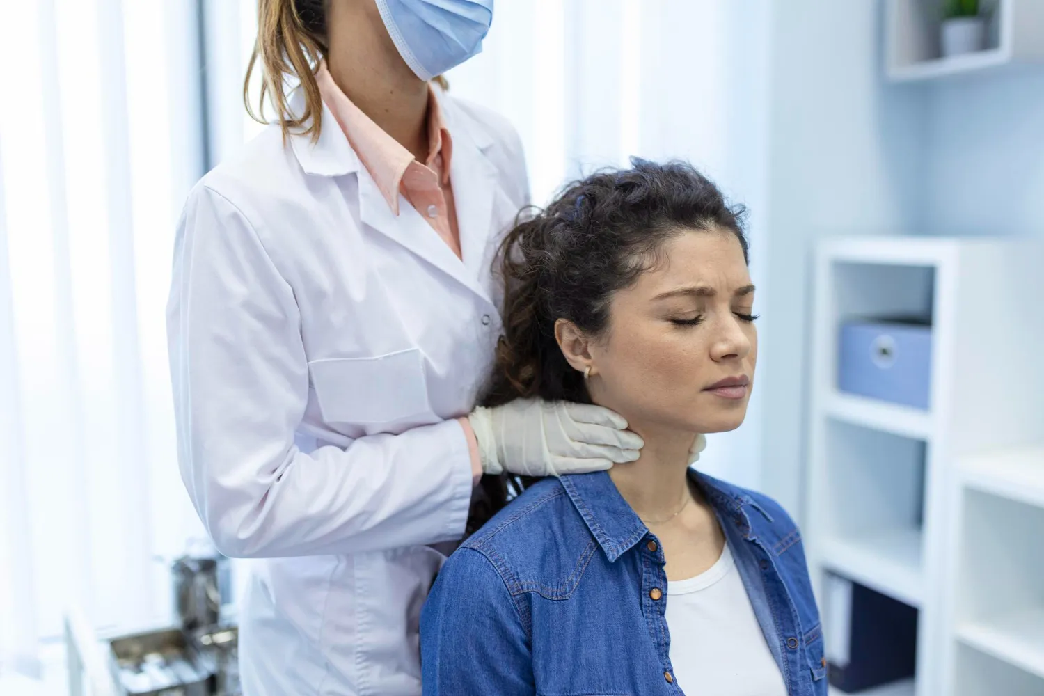 Doctor wearing gloves and mask examines patient's neck in a medical office with closed eyes.