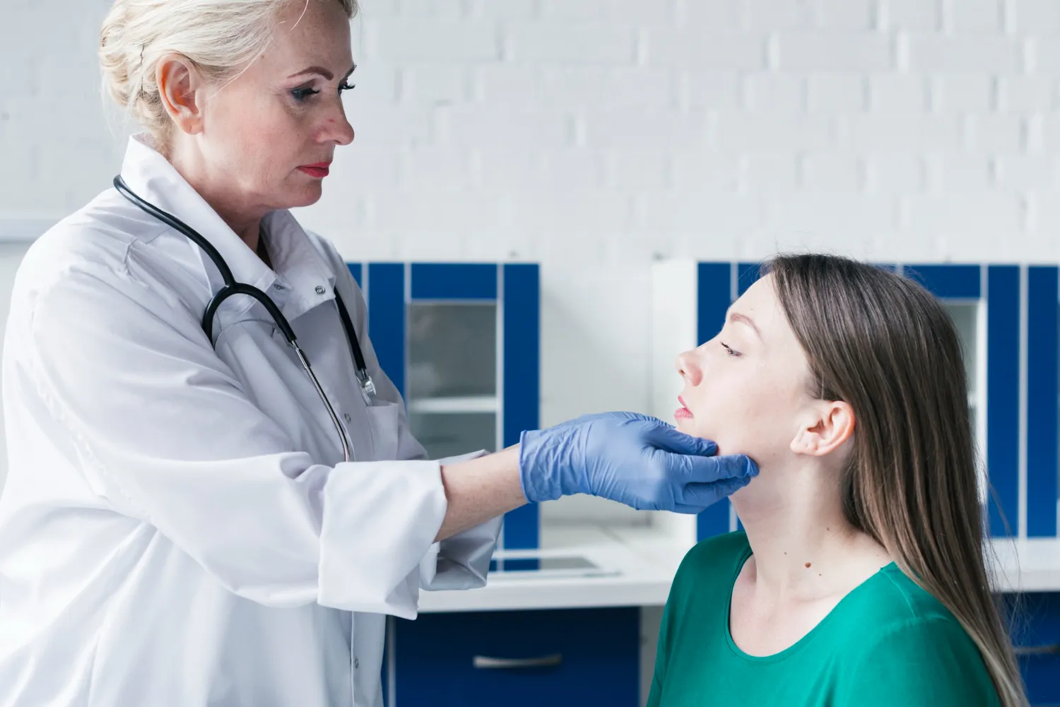 Doctor in white coat with stethoscope checking a woman's throat while wearing blue gloves.