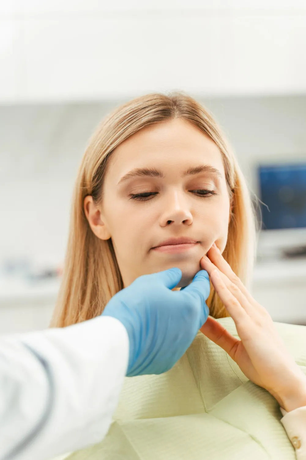 Woman touching her jaw while a doctor with blue gloves examines her chin in a clinic.