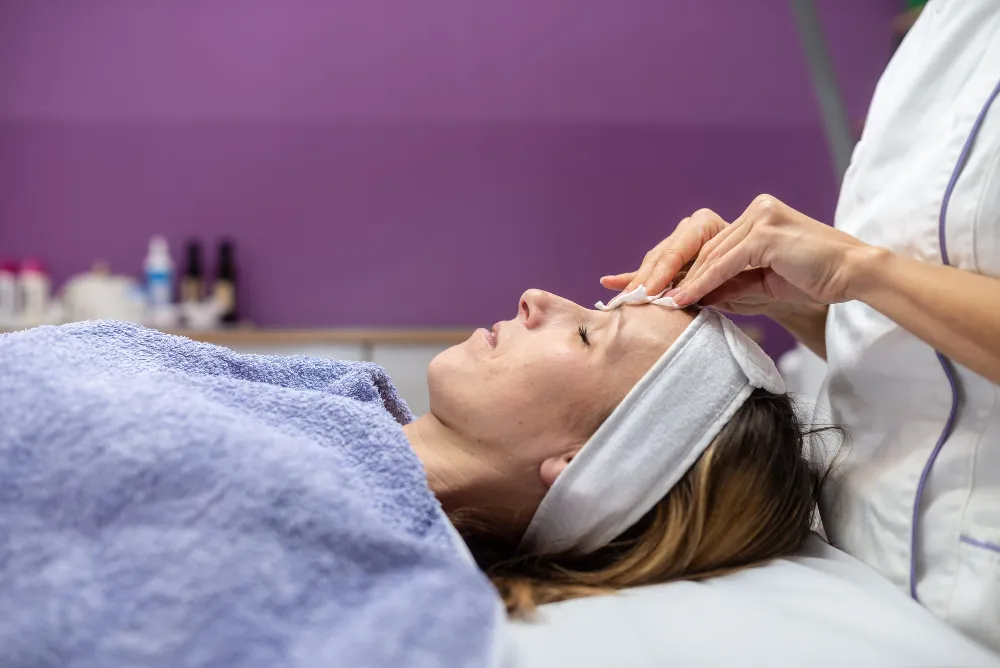 Woman receiving a facial treatment with a towel headband in a spa setting with a purple wall background.
