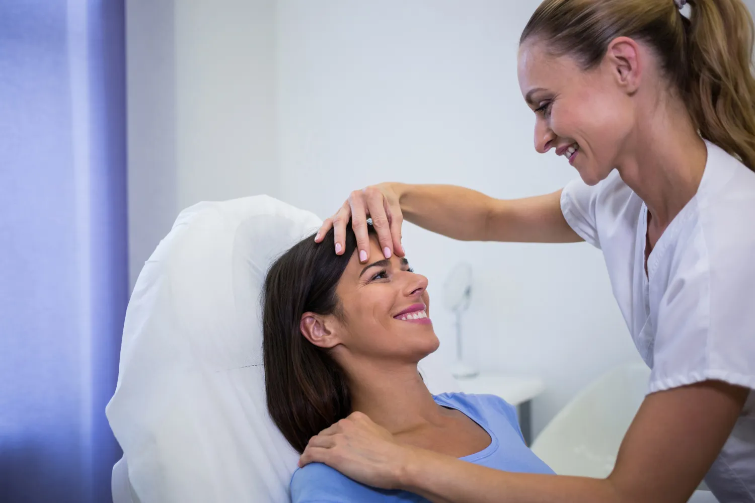 A healthcare professional gently examining a smiling woman's forehead in a clinical setting.