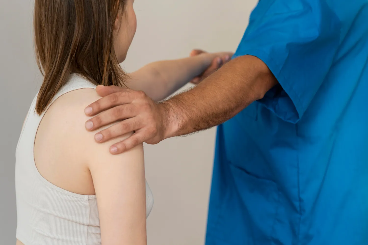 Doctor in blue scrubs examining a patient's shoulder and arm, with supportive hand on shoulder.