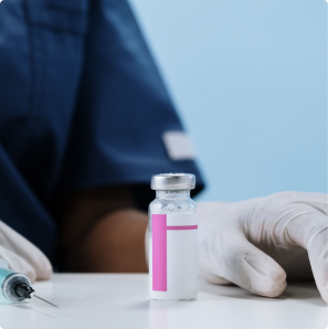 Medical injection vial and syringe prepared on a clinical table beside a healthcare professional wearing gloves.
