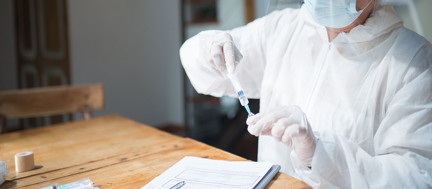 Healthcare professional wearing protective equipment preparing a syringe and medication at a clinical worktable.