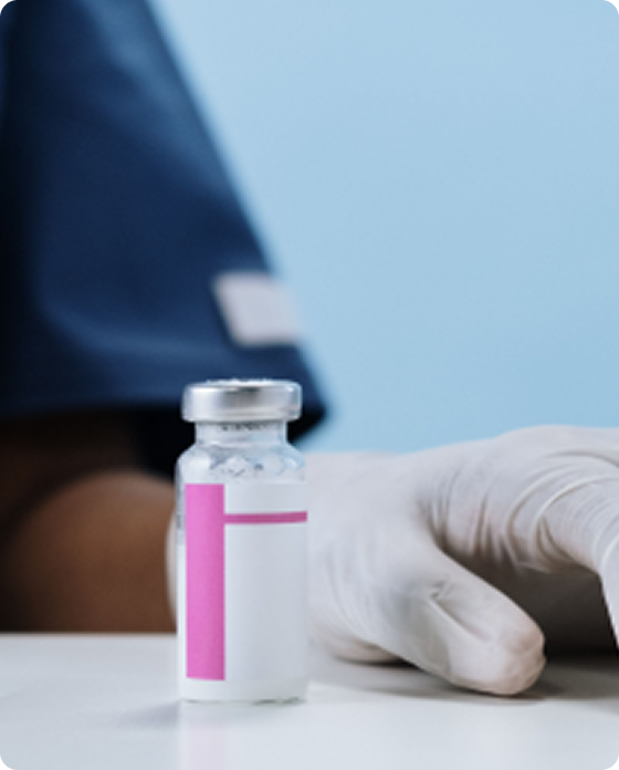 Medical injection vial and syringe on a clinical table beside a healthcare professional wearing gloves.