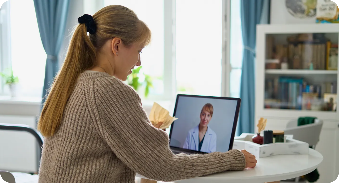 Woman with ponytail in cozy sweater video chatting with doctor on laptop at home table.