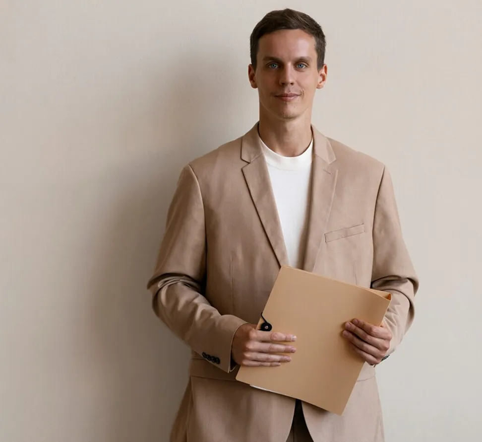 Man wearing beige suit holding a matching beige folder in front of a light beige wall.
