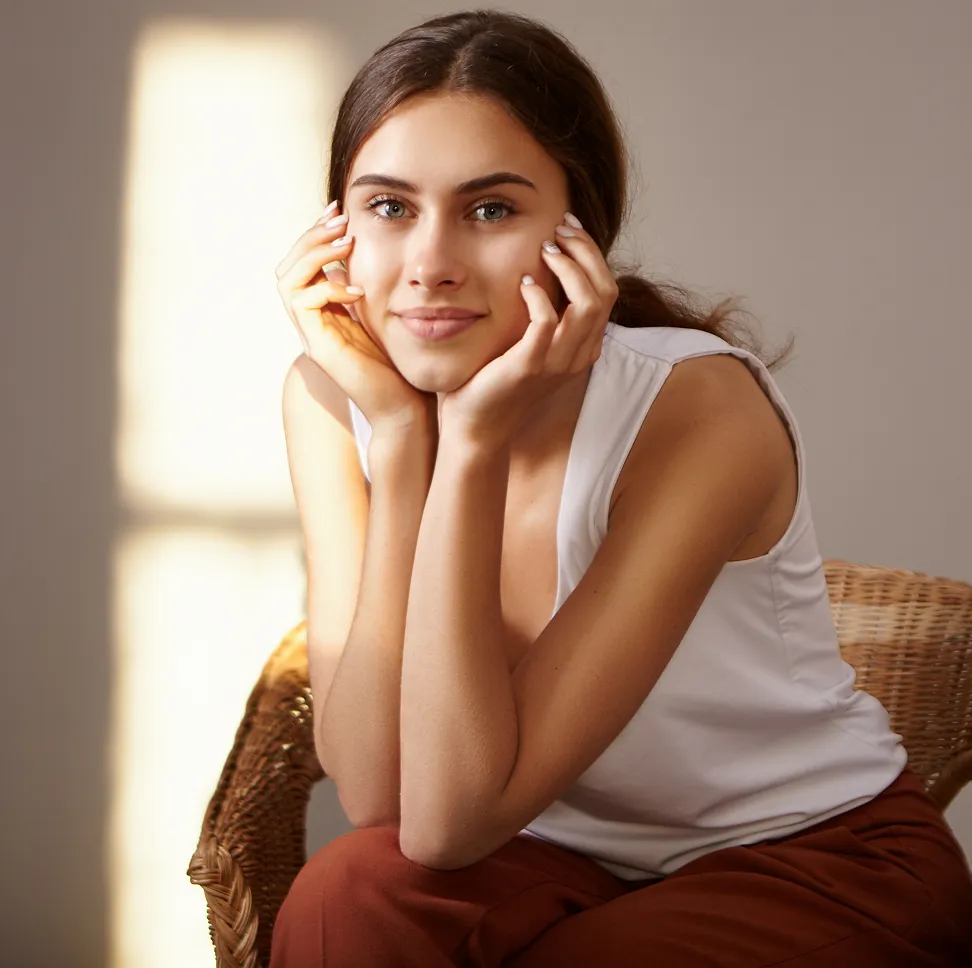 Young woman with brown hair and green eyes wearing white sleeveless top, resting her chin on hands.